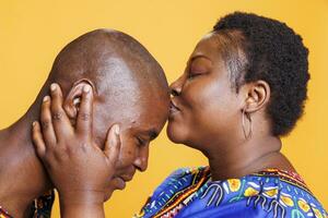 Caring wife kissing husband with closed eyes in forehead, showing couple bonding closeup. Smiling boyfriend enjoying love sign from girlfriend on orange studio background photo