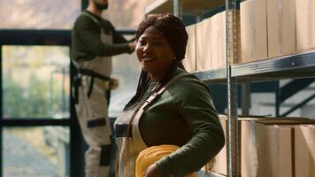 Portrait of cheerful african american warehouse technician in protective uniform dealing with packaged goods in cardboard boxes ready to be shipped, happy to work in professional facility photo