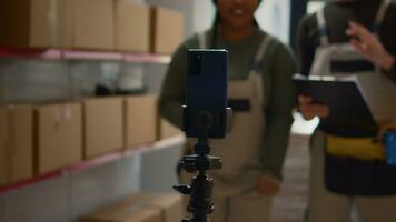 Warehouse logistics coordinator and colleague using smartphone placed on tripod to make training video for interns. Employees film themselves in fulfillment center showing trainees how to seal boxes photo