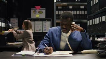 Focus on african american policeman answering phone call and taking important notes on case. In well-equipped office, expert male detective utilizes telephone to acquire evidence and analyze clues. photo