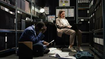 In office space, african american male cop and caucasian female inspector with laptop, examine evidence. Worn-out law officer rests on chair while policeman analyzes statements and clues on ground. photo