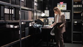 Caucasian policewoman conducts investigative legal work with confidential papers, working on a case. Examining witness statements on laptop, detective cross-references with files from archive shelves. photo
