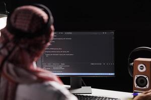 Muslim man sits at a computer and programs, exhibiting the app development process. Over-the-shoulder image of a computer monitor displaying code created by an Arab man dressed traditionally. photo