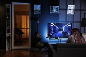 Rear-view shot of Muslim man seated on the sofa engrossed in playing video games. Image shows a Middle Eastern man dressed in traditional clothing enjoying online entertainment from the television. photo