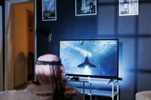 Back-view shot of gamer sitting on sofa and playing space shooter video games with controller. Man streaming online videogames for e-sport tournament in room with blue neon lights. photo