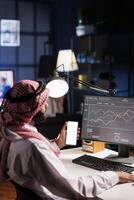 Muslim man sits at office desk, holding mobile device with blank white screen. Arab guy vertically grasping smartphone displaying isolated mockup template. Ideal for advertising or software templates. photo