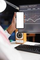 Selective focus of a guy hand holding a cellphone in front of a desktop computer with an isolated copyspace white screen. Arab showing a mobile device with a blank chromakey mockup template. photo