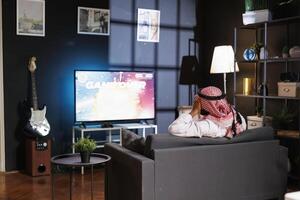 Arabic man controls the digital display while submerged in cyberspace after experiencing defeat in a virtual reality game. Seated at the sofa he has fun and is entertained with a wireless controller. photo