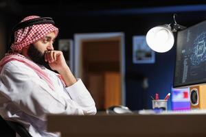 An Arab man in traditional attire works at a computer focused on coding, ensures cybersecurity and monitors the network. This image showcases the integration of technology and culture. photo