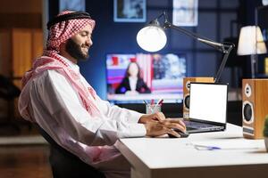 Middle Eastern man using his digital laptop that displays a blank chromakey template. A dedicated Muslim guy is utilizing his personal computer with an isolated mockup white screen. photo