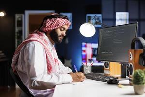 Muslim man sits at a computer and programs, exhibiting the app development process. Side-view image of a computer monitor displaying algorithms created by an Arab guy dressed traditionally. photo