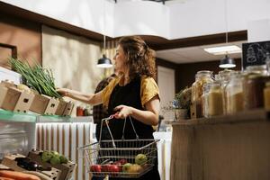 Woman in low carbon footprint zero waste store interested in purchasing food with high nutritional value. Client fills shopping basket with pesticides free produce in local neighborhood shop photo