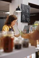 Storekeeper at zero waste supermarket checkout counter using bar code scanner to process customer transaction. Seller in sustainable local neighborhood shop scanning client food items photo