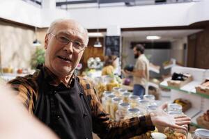 Old zero waste shop entrepreneur using selfie camera on mobile phone to promote sustainable vegan products online. Octogenarian bio supermarket owner making marketing internet content photo