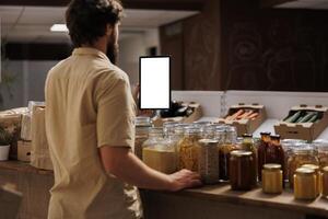 Client in zero waste shop using mockup tablet to videocall his girlfriend, buying her vegan food. Man with isolated screen device phone calls partner over internet in local supermarket photo