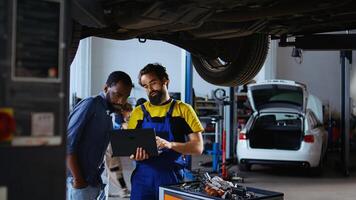Trained mechanic and customer standing underneath car in garage using laptop to order new parts after finding defects. Employee assisting african american client by looking for components online photo