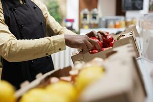 A modern supermarket owner arranges eco friendly products on a shelf. Selective focus on red peppers being organized by an african american person, highlighting local and sustainable options. photo