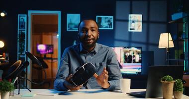 Tech content creator filming technology review of newly released gaming VR glasses for audience in neon lit home studio. BIPOC internet star showing virtual reality headset to subscribers photo