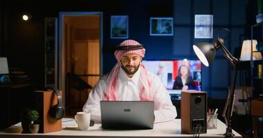 Muslim teleworker remotely working in dimly lit apartment, enjoying fresh cup of coffee. Middle Eastern worker at home typing on notebook device, sending emails and drinking hot beverage photo