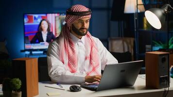 Muslim entrepreneur working from home, using laptop and writing notes. Middle Eastern man in personal apartment office answering emails and transcribing important info on notepad photo