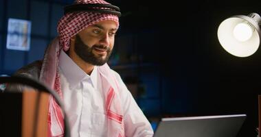 Muslim teleworker remotely working in dimly lit apartment composing emails on laptop. Middle Eastern worker at home typing business data on notebook device, wearing traditional attire, close up photo