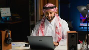 Arab businessman typing on laptop, surfing the net while at living room desk. Middle Eastern man conversing through email in professional business environment, focused on solving various tasks photo
