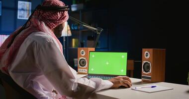 Muslim man scrolling on green screen laptop, checking social media feed. Self employed person surfing the internet from chroma key device in apartment office with TV as background noise photo