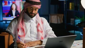 Arab entrepreneur working at home office desk on laptop, browsing the internet and writing in notebook. Muslim man using pen, notepad and digital device while teleworking photo