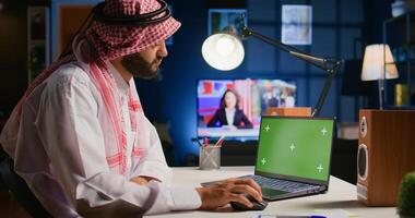 Muslim man scrolling on isolated screen laptop, checking social media posts. Self employed person browsing internet websites from chroma key device in apartment office with TV as background noise photo