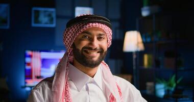 Portrait of happy arabic teleworker sending job emails in stylish apartment. Cheerful muslim employee remotely working, typing data on laptop with opened tv as background noise, close up shot photo