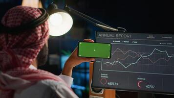 Remote worker holding green screen smartphone in landscape mode while looking at business annual report graphs on computer. Middle Eastern freelancer with mockup phone in hand performing tasks photo