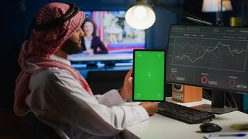 Remote worker at home holds chroma key tablet while looking at business annual report graphs on computer. Arab freelancer with mockup digital device in hand performing company tasks photo