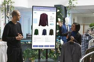 Asian woman holding a suit while assisting young man at the digital kiosk board with clothing prices. Female retail employee showing different clothes and merchandise to caucasian male client. photo