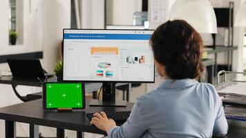 Businesswoman works at desk with greenscreen presented on modern tablet, looking at isolated copyspace template while she analyzes revenue data on pc. Worker uses chromakey display. Handheld shot. photo