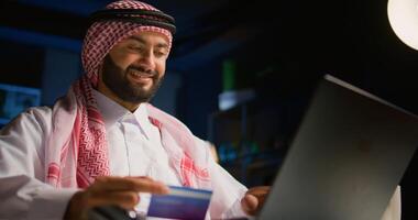 Cheerful arab man doing internet shopping, typing credit card information on computer. Smiling Middle Eastern person adding payment method on website, paying for online groceries order photo