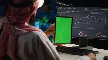 Remote worker holding isolated screen tablet while looking at business annual report graphs on computer. Middle Eastern freelancer with mockup digital device in hand performing tasks photo