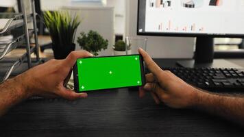 pov de trabajador sostiene teléfono con pantalla verde monitor a escritorio, trabajando con aislado Bosquejo modelo a puesto de trabajo. joven empleado mirando a móvil teléfono pantalla con clave de croma foto