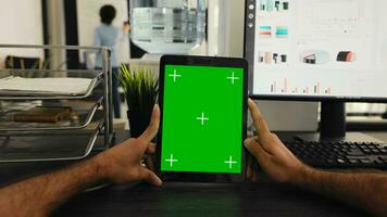 POV of businessman holding tablet with greenscreen template, looking at chromakey display and sitting coworking space desk. Person examining isolated mockup copyspace on gadget screen. photo