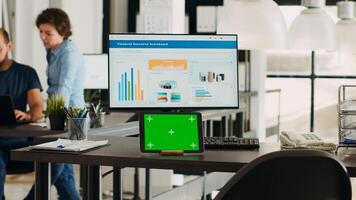 Desk with tablet showing greenscreen, empty workstation in creative agency coworking space. Modern gadget running isolated chromakey display in small business workplace, mockup screen. photo