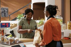 Female consumer paying with credit card at checkout counter, purchasing organic pantry staples at local supermarket. African american women at cashier desk using pos machine for seamless transactions. photo