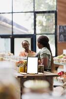 blanco pantalla en tableta muestra un anuncio modelo promoviendo fresco, orgánico y sostenible comida productos en un Respetuoso del medio ambiente mercado. vendedor habla con cliente mientras dispositivo muestra blanco Bosquejo. foto