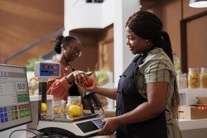 Female vendor receiving locally grown produce from young consumer for measuring weight at checkout counter. African american vendor using digital scale to weigh fresh products from customer. photo