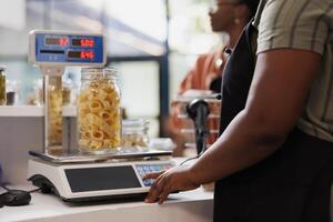 Detailed shot of seller utilizing digital scale to determine weight of container filled with organic produce. Closeup of vendor weighing jar of fresh pasta, with client patiently waiting at counter. photo