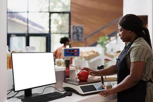 Vendor weighing organic sustainable produce on digital scale while computer displays isolated mockup template for bio food market promotion. Desktop pc at checkout counter showing white screen. photo