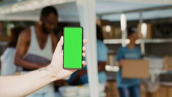 Focus on a male hand vertically holding mobile phone displaying isolated chromakey template at outdoor food bank. Close-up of caucasian person grasping cellphone with green screen. photo