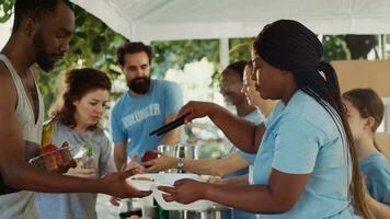 Side-view of young volunteers serving meals to those in need, helping homeless people and refugees. Poor and hungry individuals getting free food and nourishments at an outdoor food bank. Tripod shot. photo