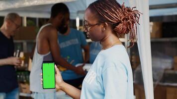 Close-up of african american lady vertically grasping mobile device having blank copyspace chromakey template. During charitable food bank black woman volunteer holds cellphone with green screen. photo