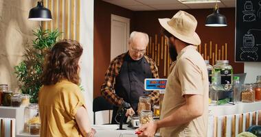 Older vendor assisting clients in zero waste shop with ethical sourced food items, inviting them at checkout. Couple in local store buying pantry staples bulk products, tracking shot photo