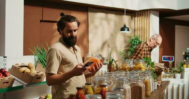 Man in zero waste shop buying vegetables, using phone to check products online before making purchase decision. Vegan client taking photo of food in local store, verifying it on specialty website