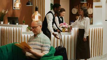 Older guest arriving on vacation at hotel with room reservation, bellhop offering concierge services carrying baggage for tourist. Receptionist doing check in with woman at front desk. Tripod shot. photo